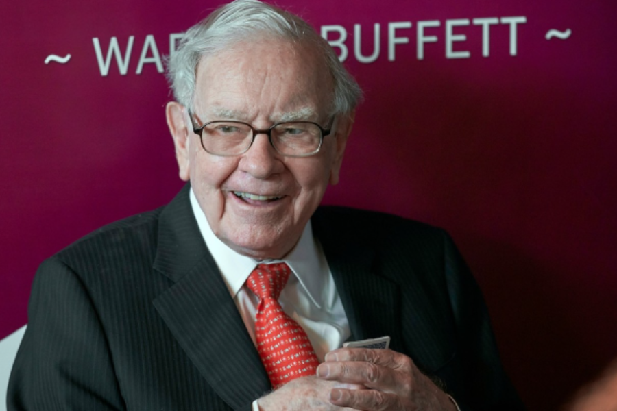 An elderly man with white hair and glasses smiles warmly while seated in front of a maroon backdrop that reads "~ WARREN BUFFETT ~". He wears a dark pinstripe suit, a white shirt, and a red patterned tie, with his hands gently clasped in front of him.