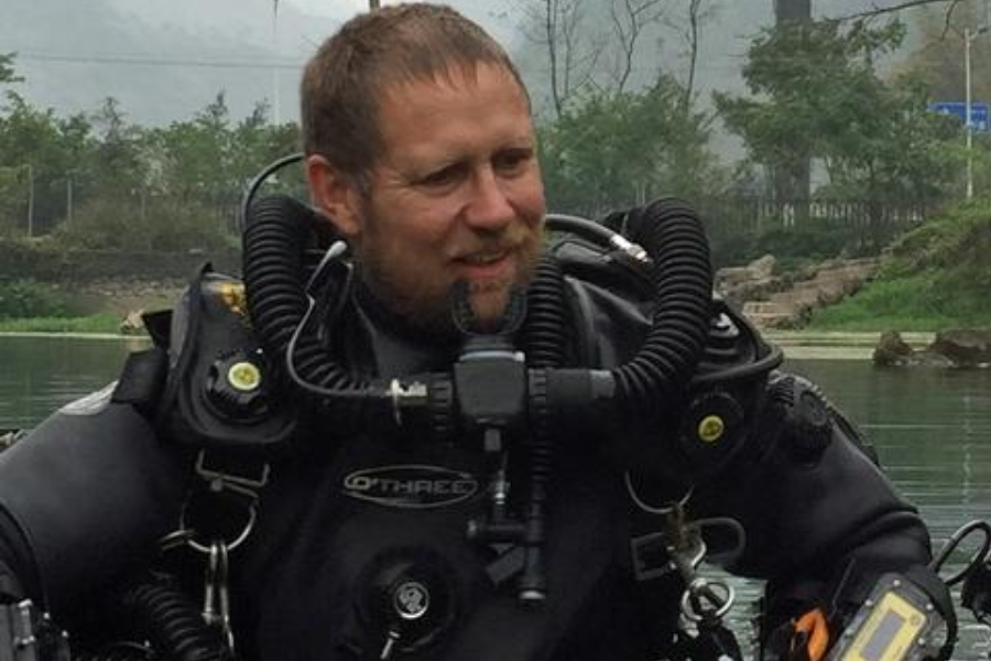 A smiling cave diver in a heavy-duty drysuit and advanced rebreather system prepares near a calm body of water, surrounded by greenery and misty hills. The diver’s suit is marked “O’THREE,” and he appears ready for a technical dive, likely related to a rescue or training mission.