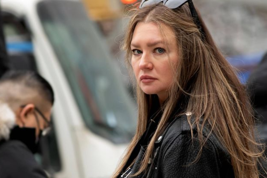 A woman with long, light brown hair and a serious expression looks over her shoulder while standing outdoors. She wears a black leather jacket and sunglasses perched on her head, with blurred city elements and pedestrians in the background.