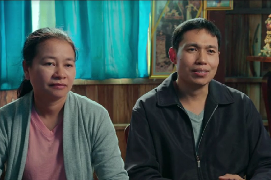 A woman in a light gray cardigan and a man in a dark jacket sit side by side in a warmly lit room with turquoise curtains and framed photos in the background. Both appear calm and thoughtful, as if sharing their story or giving an interview about a significant experience.
