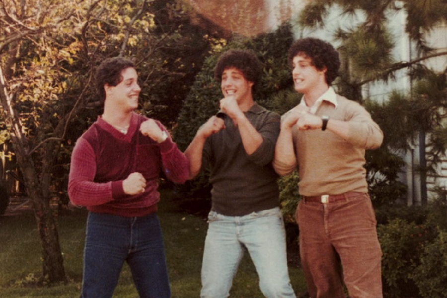 Three young men with curly hair pose playfully in a mock boxing stance on a sunny day, all smiling and wearing vintage sweaters and jeans. The scene takes place in a grassy yard with trees and shrubs in the background.