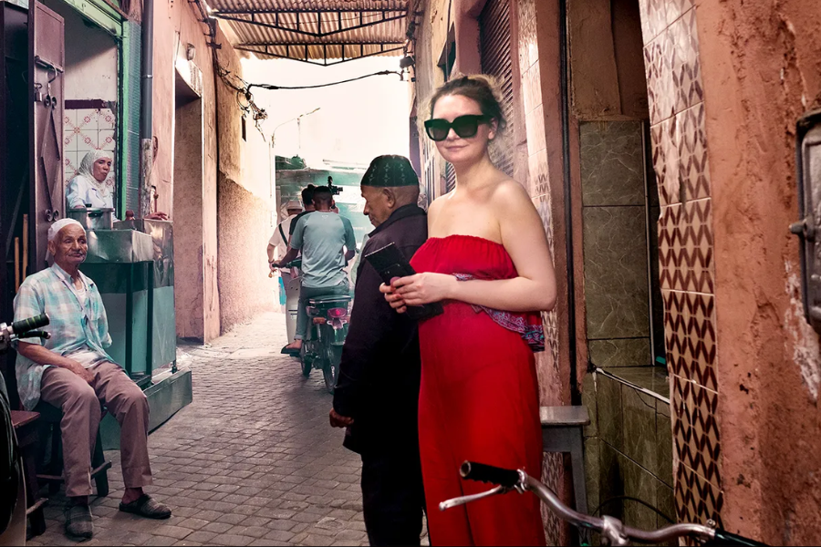A woman in a strapless red dress and oversized sunglasses stands confidently in a narrow alleyway lined with patterned tiles and small shops. She holds a phone and smiles slightly, while locals sit or ride scooters through the vibrant, sunlit passageway behind her.