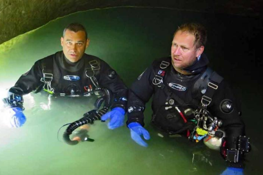Two cave divers in full black drysuits and blue gloves stand in waist-deep water inside a dimly lit cave. Both men wear technical diving gear with rebreathers and tanks, appearing focused and ready for an underwater rescue or exploration operation.