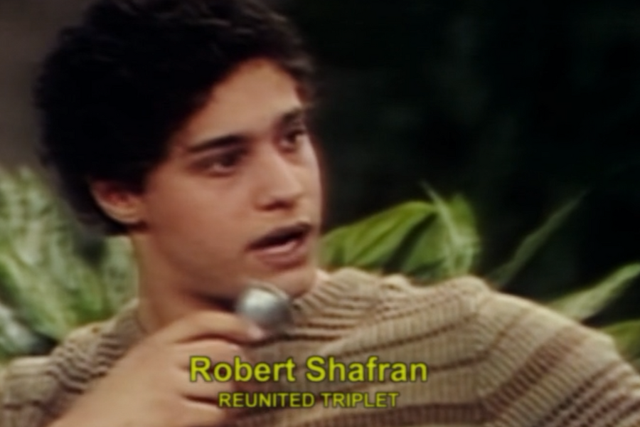 Young man speaking into a microphone on a television show, identified by on-screen text as "Robert Shafran, Reunited Triplet." He wears a beige striped sweater and sits in front of green leafy plants.