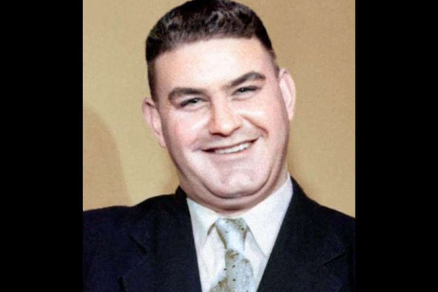 Colorized portrait of a smiling man in a dark suit and patterned tie, posed against a neutral beige background. He has neatly styled dark hair and a cheerful expression.
