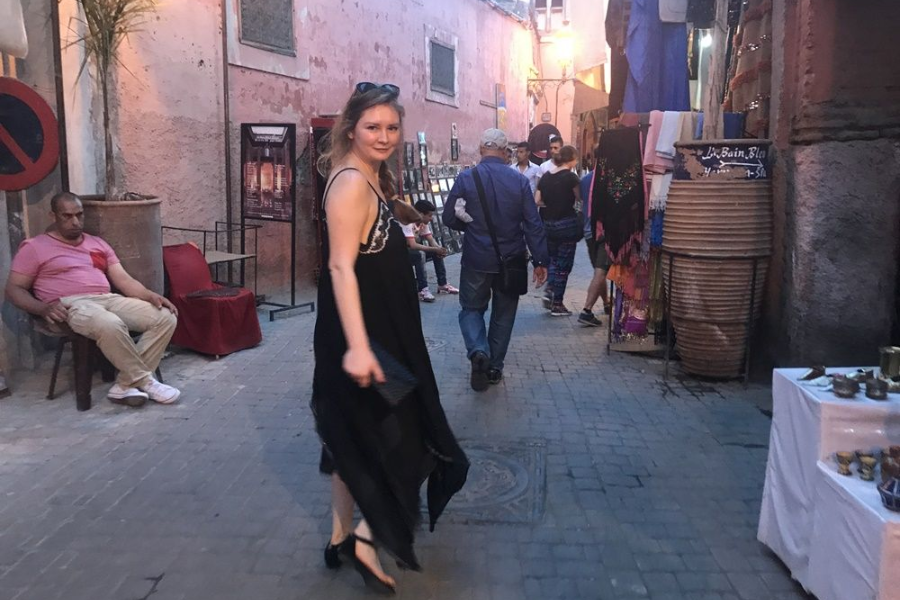 A woman in a flowing black dress with lace trim glances back at the camera while walking through a narrow, bustling market street lined with textiles, lanterns, and souvenir shops. The street is lit with warm tones as people browse, and a man in a pink shirt sits relaxed on the left side.