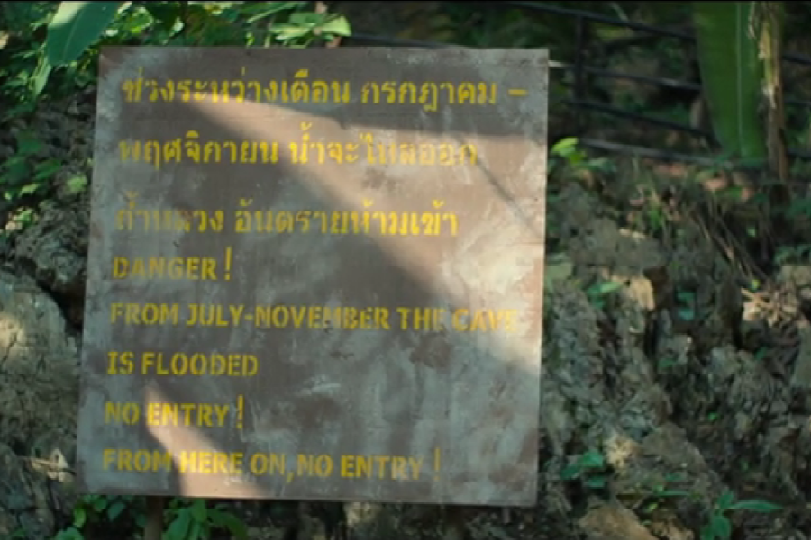 A warning sign posted in front of a rocky, wooded area, with text in both Thai and English. The English portion reads: “DANGER! FROM JULY–NOVEMBER THE CAVE IS FLOODED. NO ENTRY! FROM HERE ON, NO ENTRY!” indicating hazardous cave conditions during monsoon season.
