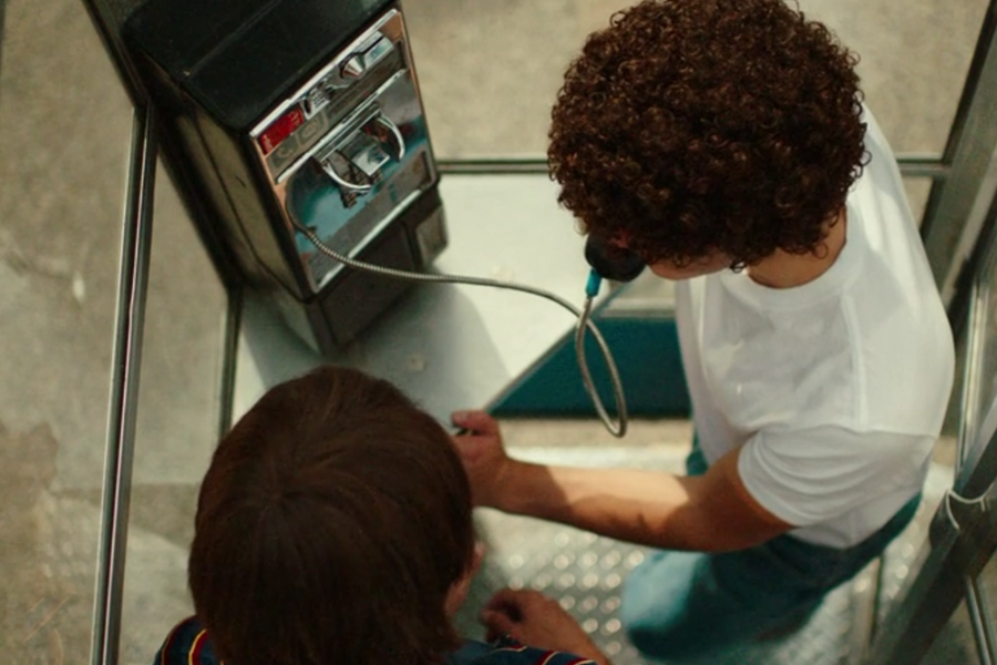 Overhead view of two people inside a phone booth, with one person in a white shirt using a corded payphone while the other stands close beside them. The scene captures a retro vibe, with the shiny metal phone and booth details visible.