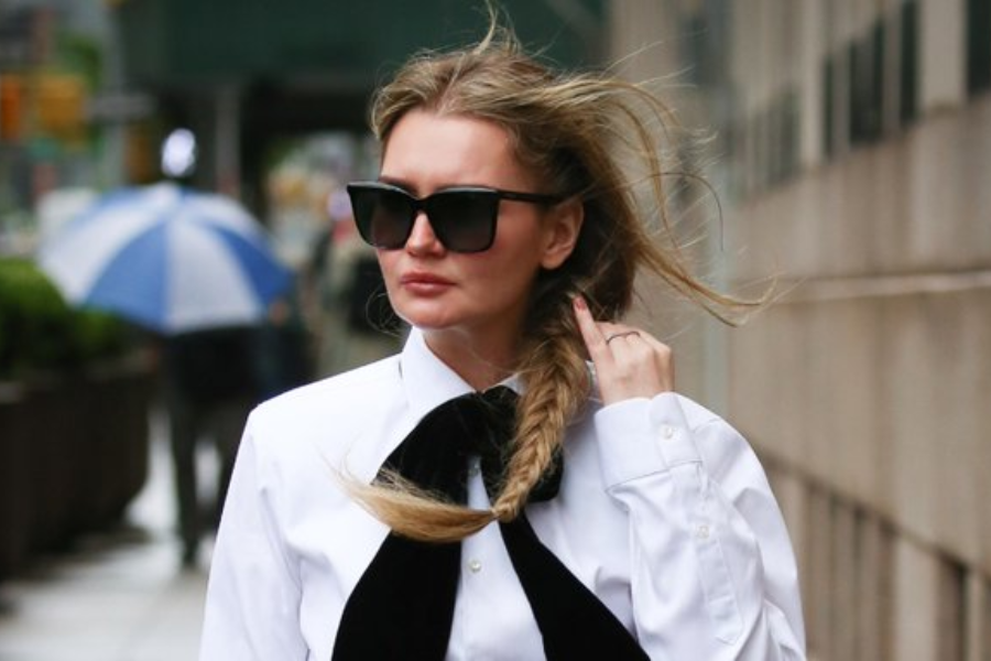 A woman walks on a city sidewalk wearing a crisp white button-up shirt with a large black velvet bowtie, her hair in a side fishtail braid blowing in the wind. She wears oversized black sunglasses and a serious expression, with a blue-and-white umbrella visible in the blurred background.