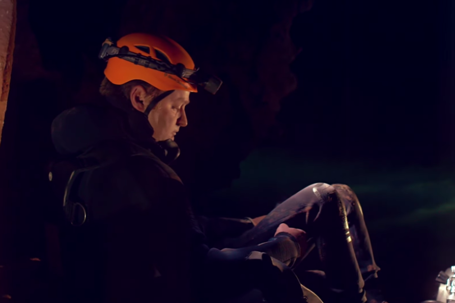 A rescue diver in a black wetsuit and orange helmet with a headlamp sits alone inside a dark cave, resting against a rocky wall. His body language and dim lighting suggest fatigue and contemplation amid a demanding cave operation.