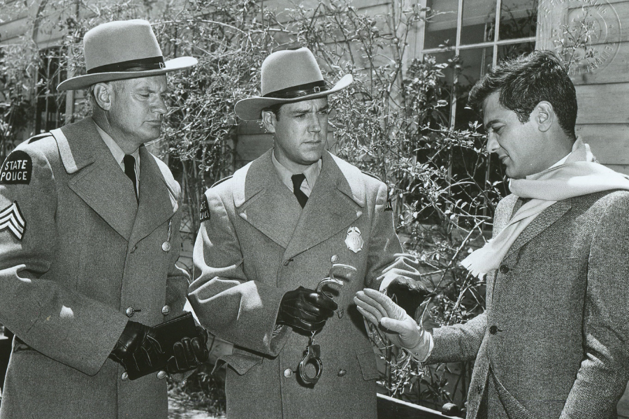 Black and white film scene showing two uniformed state police officers preparing to arrest a well-dressed man in a scarf and overcoat. One officer holds handcuffs while the man extends his hands calmly, standing in front of a building with climbing plants.