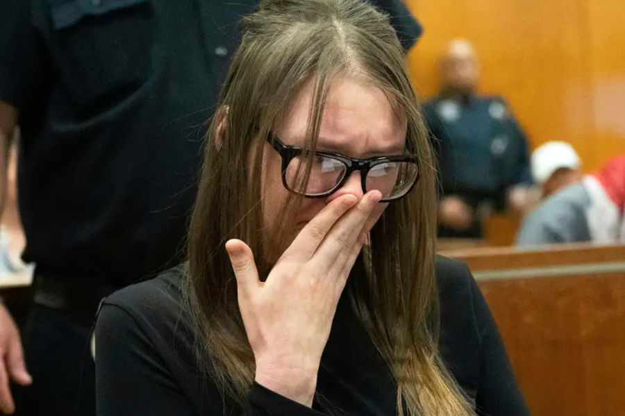 A woman with long brown hair and large black glasses wipes away tears in a courtroom, visibly emotional with her hand covering part of her face. She wears a black outfit, and uniformed officers and other people can be seen in the blurred background.