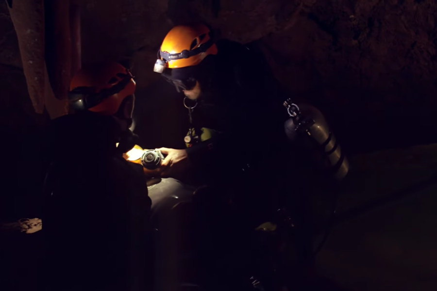 Two rescue divers wearing orange helmets and black wetsuits work together in a dark cave, illuminated only by their headlamps. One diver appears to be adjusting or inspecting a piece of equipment while the other holds it steady, highlighting their teamwork in the challenging conditions.