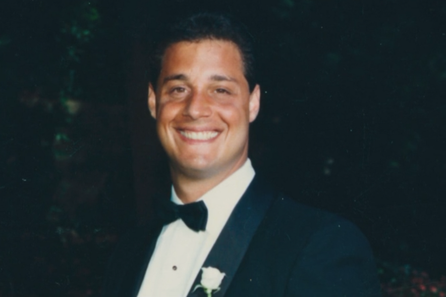 Smiling man in a tuxedo with a white rose boutonniere, posing for a formal portrait against a dark, softly blurred background. His cheerful expression suggests a celebratory occasion, possibly a wedding or special event.