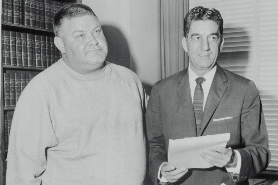 Black and white photo of a man in a plain sweatshirt standing beside a suited man holding a document. They appear to be in an office or legal setting, with shelves of books and window blinds in the background.