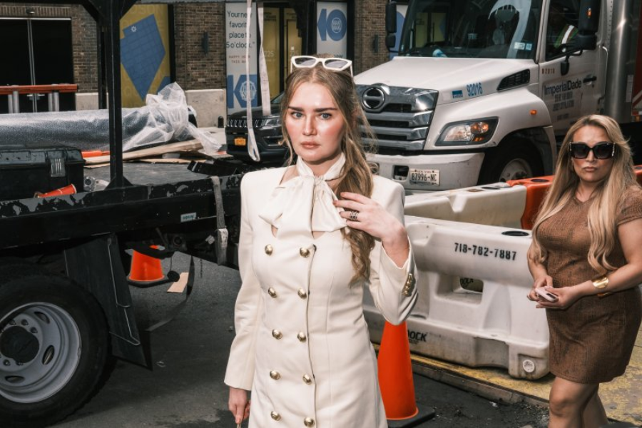 A woman in a chic cream trench dress with gold buttons and an oversized neck bow stands confidently amid a gritty construction site, with a flatbed truck and traffic cones in the background. She wears white sunglasses perched on her head, while another woman in a brown dress and sunglasses walks behind her.