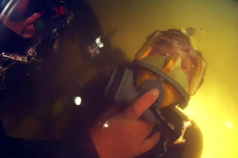 A diver gently holds the face mask of a child submerged just below the surface of yellow-tinted cave water, guiding them through a rescue. The child's eyes are closed, and the full-face mask provides breathing support during the tense underwater extraction.