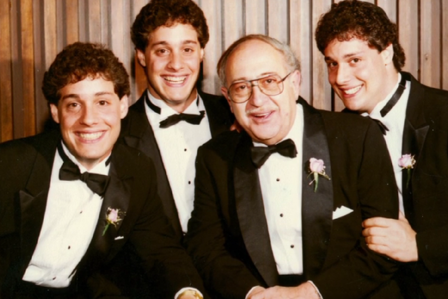 Three young men with matching curly hair and tuxedos pose cheerfully alongside an older man, also in a tuxedo, in front of a wood-paneled wall. All four wear boutonnières, suggesting a formal event or celebration.