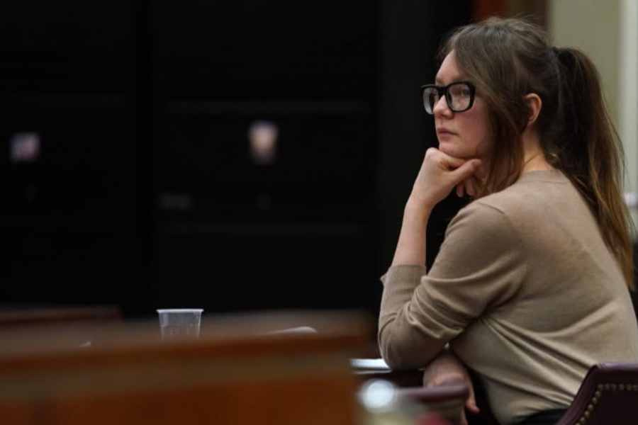 A woman with a ponytail and black-framed glasses sits in a courtroom with her chin resting on her hand, appearing deep in thought. She wears a beige sweater and looks toward the front of the room, with a plastic cup and papers faintly visible on the table.