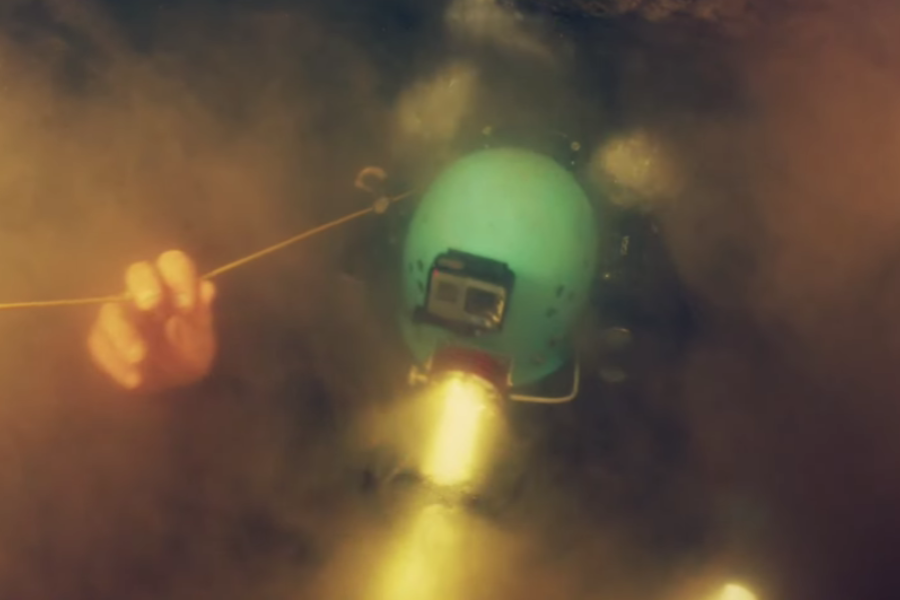 An underwater cave diver, wearing a teal helmet equipped with a mounted action camera and headlamp, navigates through murky water while gripping a guide line. The diver’s beam cuts through the silt-filled darkness, emphasizing the low visibility and difficulty of the cave rescue.