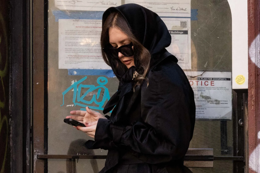 A woman dressed in an all-black outfit with a dramatic hood and oversized sunglasses looks down at her phone while standing in front of a door covered with official notices and graffiti. Her attire is strikingly formal against the gritty urban backdrop.