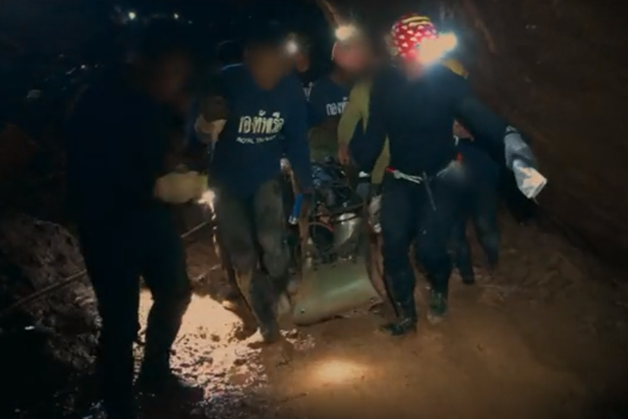 Rescuers in a dark, muddy cave work together to carry a stretcher, possibly transporting one of the trapped individuals. They wear headlamps and navy gear, with “Royal Thai Navy” visible on some shirts, highlighting the intense and coordinated effort of the cave rescue mission.