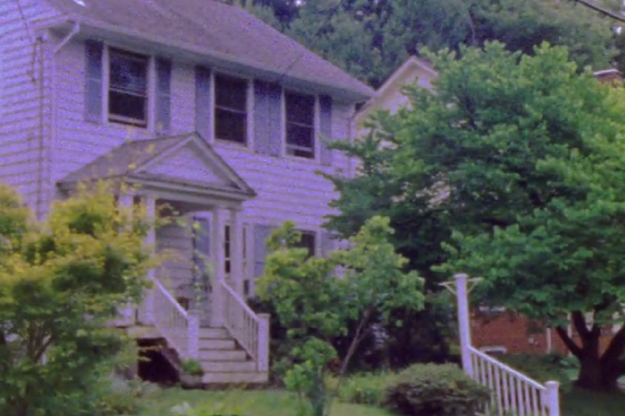 Vintage-looking photo of a two-story suburban house with white siding, black shutters, and a small front porch with railings, surrounded by lush green trees and bushes.