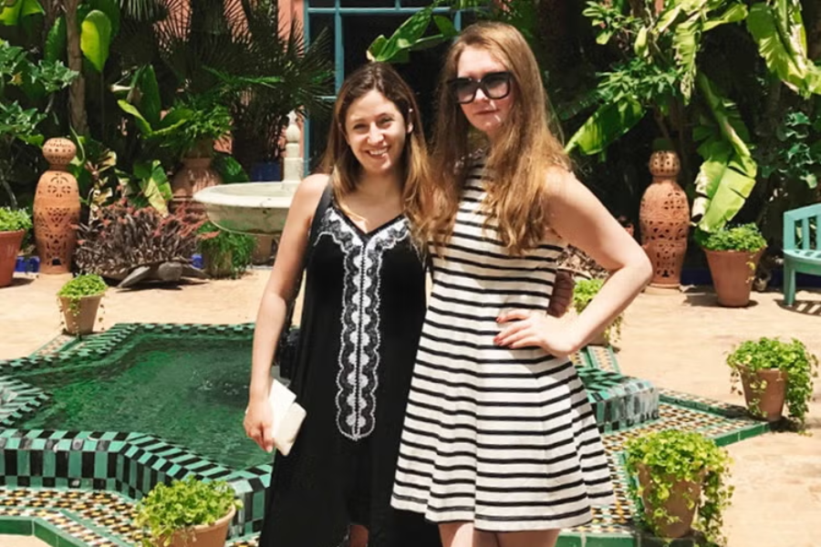 Two women pose in front of a vibrant tiled fountain surrounded by lush greenery and terracotta pots in a courtyard. One wears a black dress with white embroidery and holds a clutch, while the other wears a sleeveless black-and-white striped dress and oversized sunglasses, striking a confident pose.