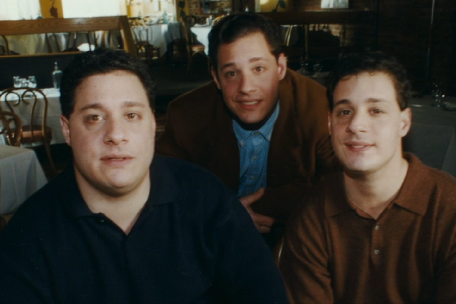 Three adult men pose closely together inside a restaurant, all with similar facial features suggesting they are siblings or triplets. The setting includes wooden chairs and white tablecloths, adding a warm, casual ambiance.