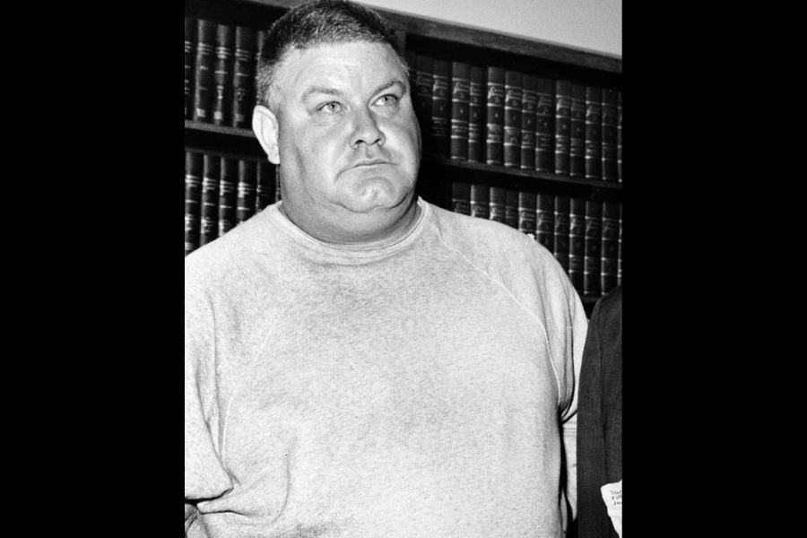 Black and white photo of a stern-looking man in a plain sweatshirt standing in front of a bookshelf filled with legal volumes. His expression is tense, and only part of another suited individual is visible beside him.
