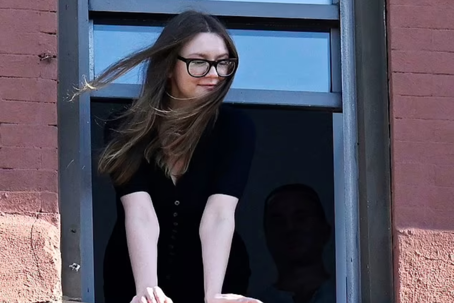 A woman with long hair and large black glasses leans out of a window, her hair blown slightly by the wind as she looks to the side with a faint smile. She wears a black buttoned dress, framed by a red brick building exterior.