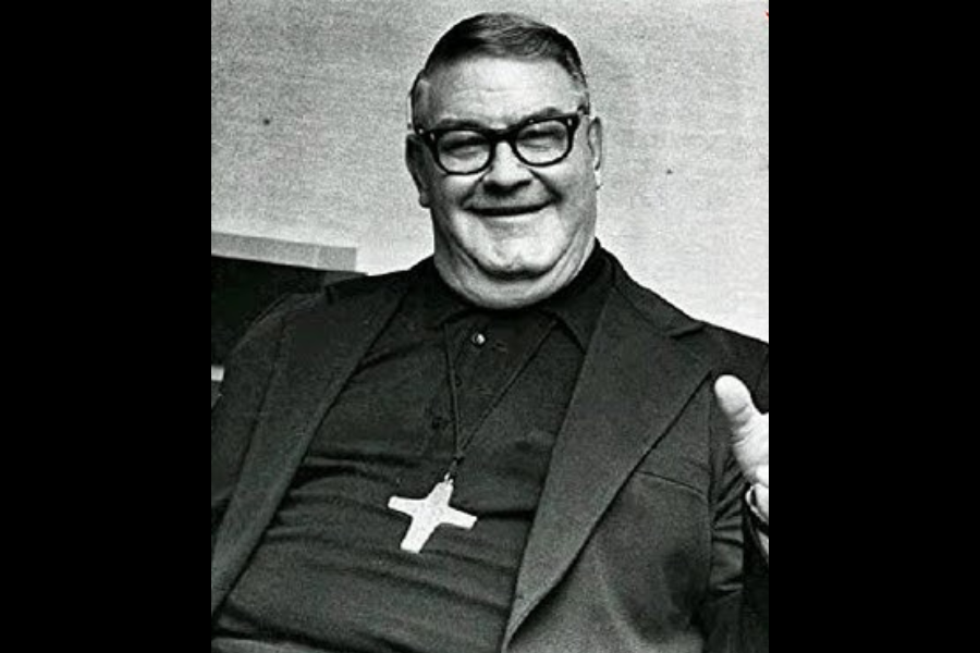 Black and white photo of a smiling man wearing glasses and clerical clothing with a large cross necklace. He is seated and gesturing with one hand, appearing relaxed and friendly.