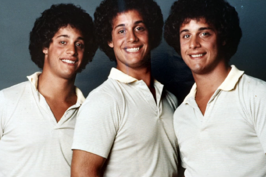 Three identical young men with curly hair stand smiling in a studio portrait, all wearing light yellow collared shirts. Their close resemblance and matching outfits highlight their identity as triplets.