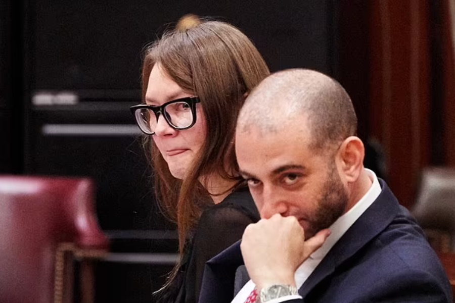 A woman in black glasses appears visibly emotional, pressing her lips together as she sits beside a man in a suit, likely her attorney, who rests his chin on his hand with a pensive expression. They are seated in a courtroom setting, with maroon leather chairs and legal documents in the background.