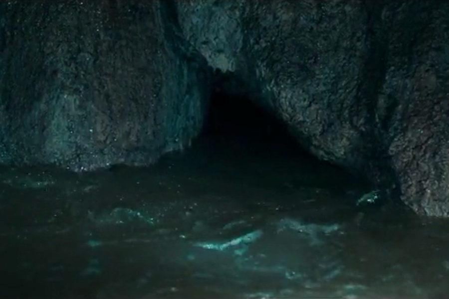 A narrow, partially submerged cave passage with dark, murky water flowing beneath rugged rock walls. The entrance to the tunnel is barely visible, emphasizing the tight and dangerous conditions rescuers must navigate during an underwater cave mission.