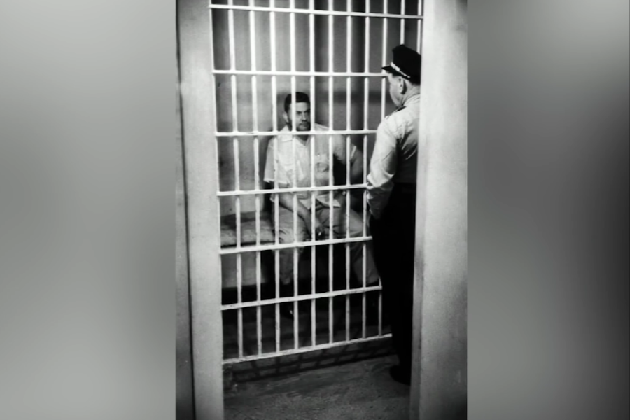 Black and white photo of a man sitting on a bed inside a jail cell, wearing a short-sleeve button-up shirt and pants. A uniformed officer stands outside the bars, appearing to speak with the inmate.