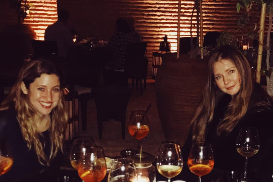 Two women sit at an elegantly dim-lit outdoor restaurant table, smiling amid several glasses filled with orange cocktails and white wine. The ambiance is warm with candlelight and backlit textured walls, creating a cozy, upscale dining atmosphere.