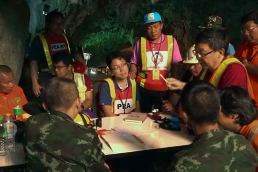 A group of rescue coordinators, including military personnel and engineers in high-visibility vests marked "PEA" and "PEANK," gather around a table in a cave to discuss plans. One man gestures with a tool while others listen intently, highlighting the intense collaboration during the cave rescue operation.