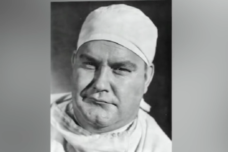 Black and white portrait of a man dressed as a surgeon, wearing a surgical cap and gown. He looks directly at the camera with a serious, focused expression.