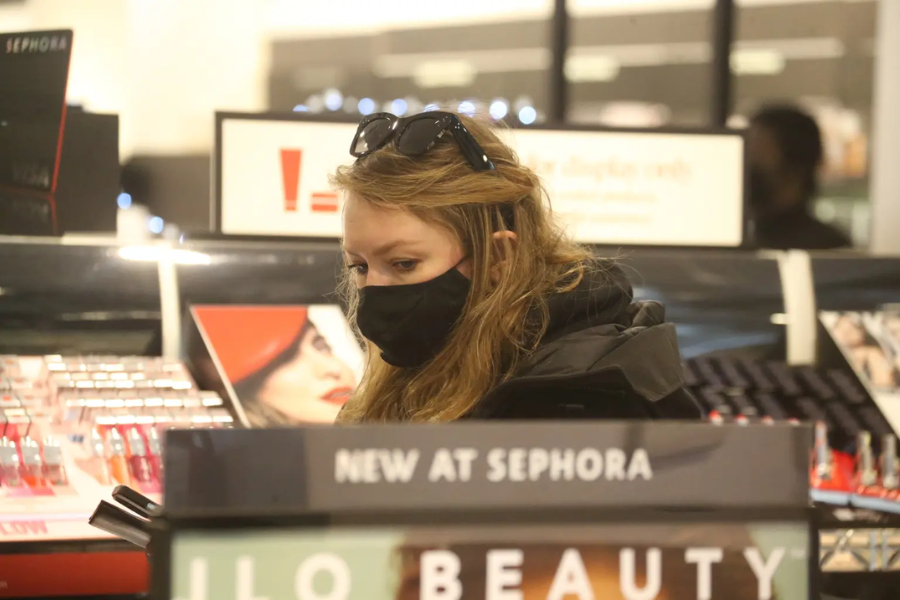 A woman wearing a black face mask and sunglasses on her head browses through a cosmetics display inside a Sephora store. She is surrounded by shelves of lip products and signs promoting new beauty launches, including a prominent "NEW AT SEPHORA" label.