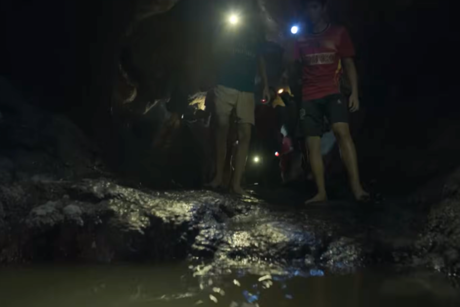 A group of barefoot boys with headlamps walk cautiously through a dark, muddy cave passage with shallow water on the floor. The dim lighting highlights the slick, rocky surface and close cave walls, creating a tense and adventurous atmosphere.
