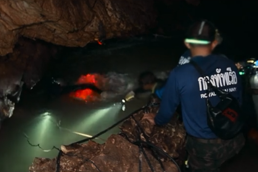 A member of the Royal Thai Navy stands at the edge of a cave pool, observing divers with flashlights submerged in murky water. The scene is dimly lit and tense, underscoring the challenges of the underwater cave rescue mission.
