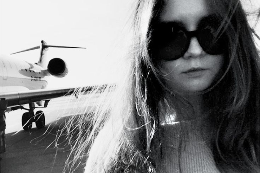 A black and white photo of a woman with long hair and dark sunglasses standing near a private jet on a tarmac. Her expression is neutral, with strands of hair blowing across her face, capturing a moody, high-fashion travel vibe.