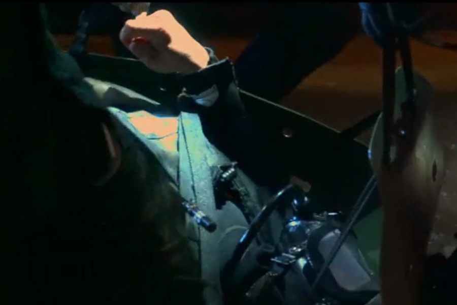 Close-up of a rescuer adjusting diving equipment attached to a stretcher or transport harness inside a dimly lit cave. The scene highlights the intricate setup and care required to safely move individuals through the underwater rescue route.