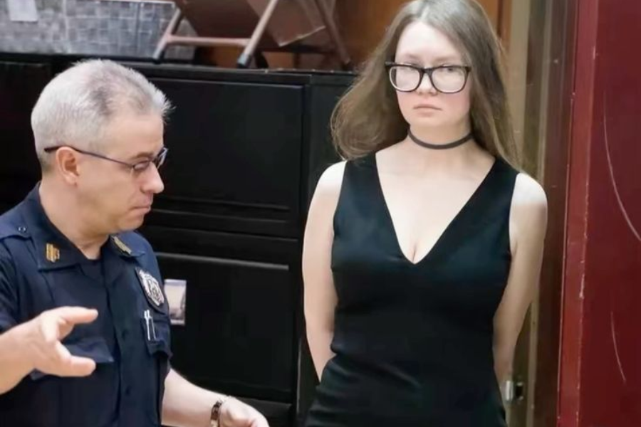 A woman with long brown hair and large black glasses stands with her hands behind her back in a courtroom, wearing a black sleeveless dress and a choker necklace. A uniformed officer beside her gestures while speaking, and her expression appears serious and composed.