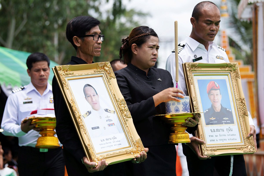 Family and military personnel solemnly hold framed portraits and ceremonial offerings during a memorial procession for two Thai Navy SEALs. The images honor those who died during the Tham Luang cave rescue mission, with one portrait showing a man in a white naval uniform and the other in a black uniform with a red beret.