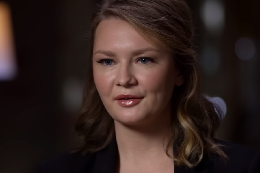A woman with softly curled, shoulder-length hair and natural makeup speaks in a dimly lit interview setting. She wears a dark blazer and gazes slightly past the camera with a composed, attentive expression.