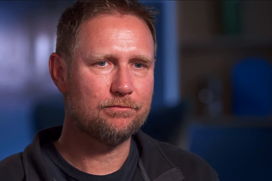 A man with short brown hair and a trimmed beard appears thoughtful and slightly emotional during an interview. He wears a dark jacket and is seated indoors in a softly lit room, suggesting he is recounting a significant and personal experience.