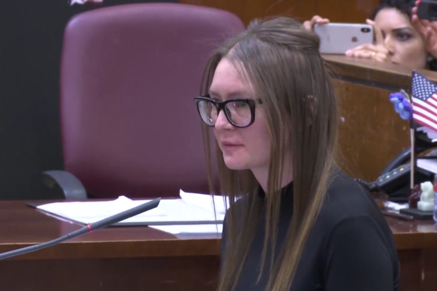 A woman with straight brown hair and large black glasses stands at a courtroom podium, facing forward with a solemn expression. Papers, a microphone, and a small American flag are visible, while people in the background capture the moment on their phones.