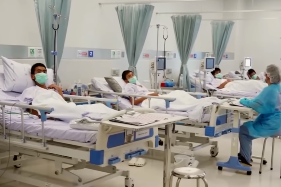 Several boys lie in hospital beds wearing face masks in a clean, well-lit medical ward, each attended by monitors and IV stands. A healthcare worker in protective gear sits at a bedside, indicating the boys are receiving care and recovery following a serious incident.